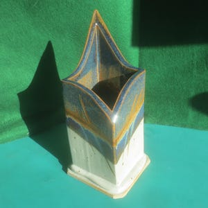 May include: A ceramic vase with a unique, angular design. The vase features a white base, transitioning to blue and gold hues at the top. The vase is approximately 8 inches tall and has a square base.