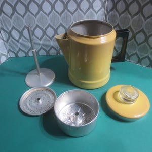 May include: A yellow metal coffee percolator with a black handle and a glass lid. The percolator is disassembled, showing the metal filter, the metal base, and the lid.