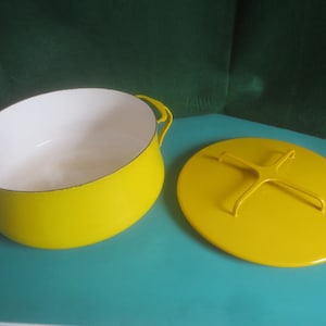 May include: A bright yellow enameled cast iron Dutch oven with a white interior and matching lid. The lid has a yellow handle. The pot has two curved handles. The Dutch oven is on a teal surface.