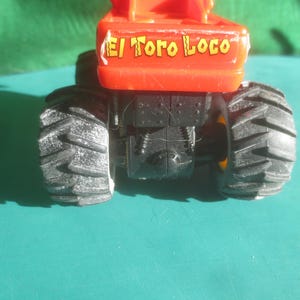 May include: A close-up of a toy monster truck with large black tires and an orange body. The text "El Toro Loco" is printed on the orange body. The truck is on a teal surface.