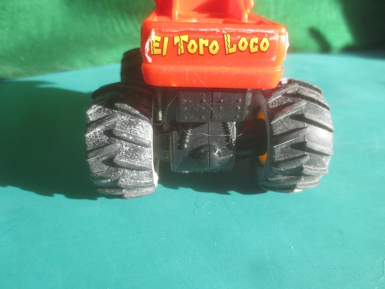 May include: A close-up of a toy monster truck with large black tires and an orange body. The text "El Toro Loco" is printed on the orange body. The truck is on a teal surface.