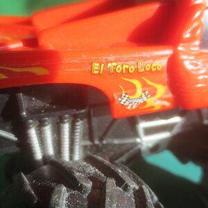 May include: Close-up of a red toy monster truck with black tires and suspension. The truck has the words "El Toro Loco" in yellow and orange flames on the side, along with a checkered flag graphic.
