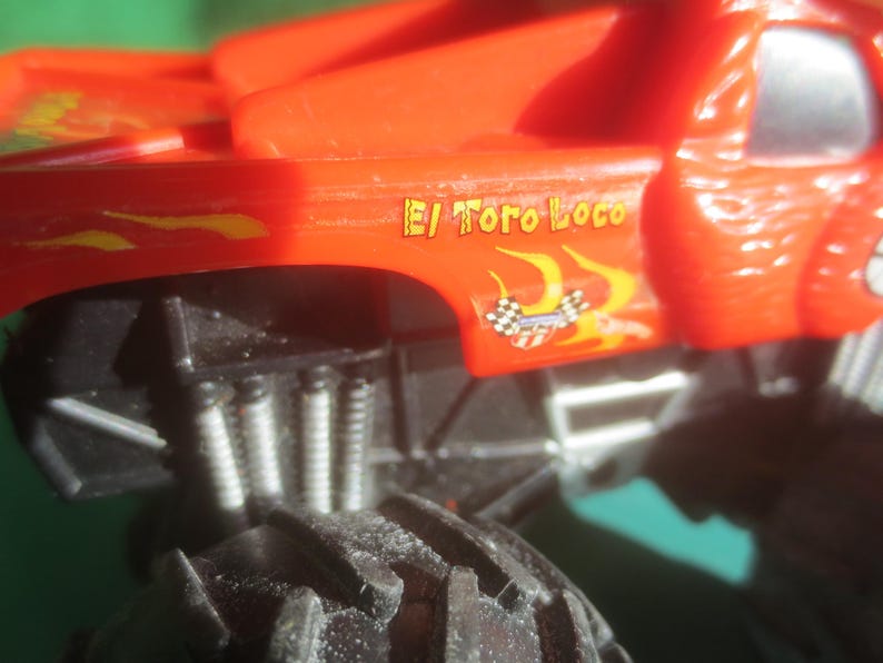 May include: Close-up of a red toy monster truck with black tires and suspension. The truck has the words "El Toro Loco" in yellow and orange flames on the side, along with a checkered flag graphic.