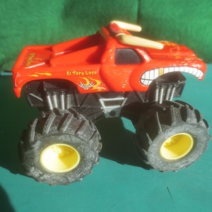 May include: A red toy monster truck with large black tires and yellow wheel centers. The truck has a bull-like design with horns and the text "El Toro Loco".