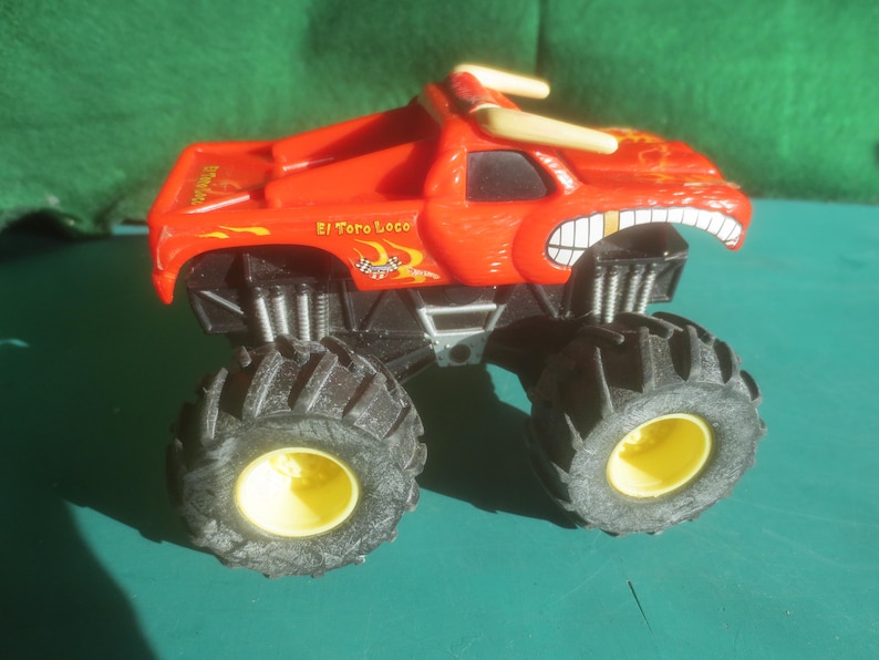 May include: A red toy monster truck with large black tires and yellow wheel centers. The truck has a bull-like design with horns and the text "El Toro Loco".