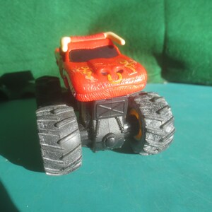 May include: A red monster truck toy with black tires and a bull-like design on the front. The truck has a black chassis and yellow accents on the wheels. The toy is made of plastic.