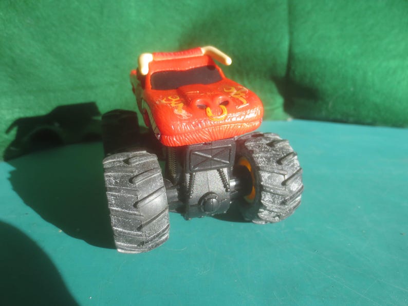 May include: A red monster truck toy with black tires and a bull-like design on the front. The truck has a black chassis and yellow accents on the wheels. The toy is made of plastic.