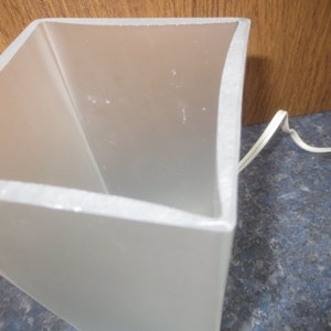 May include: A white frosted glass lampshade with a square base and a cord.