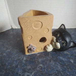 May include: A ceramic cheese-shaped planter with a black cat figurine peeking out of the side. A small grey mouse figurine is also visible on the cheese.
