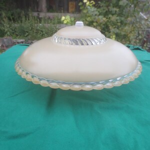 Peut inclure: Un abat-jour en verre crème vintage avec une bordure décorative en verre transparent en forme de corde. L'abat-jour a une forme arrondie en forme de soucoupe et un petit bouton blanc sur le dessus. L'abat-jour est posé sur une surface verte.