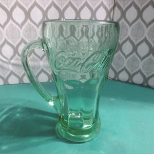 May include: A vintage green Coca-Cola glass mug with a handle. The Coca-Cola logo is embossed on the front. The mug is made of clear glass and has a classic design. The mug is on a teal surface.
