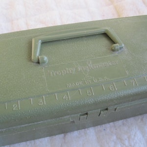 May include: A green plastic tackle box with a black handle and a ruler on the top. The box has the text "Trophy Tacklemaster" and "Made in U.S.A." printed on it.