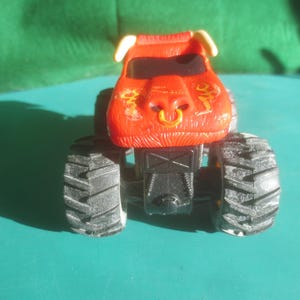 May include: A red monster truck toy with black tires. The truck has a bull-like design on the front, with yellow accents and a gold ring in the nose. The tires are large and have a rugged tread pattern.