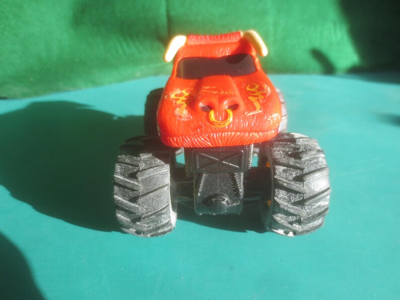 May include: A red monster truck toy with black tires. The truck has a bull-like design on the front, with yellow accents and a gold ring in the nose. The tires are large and have a rugged tread pattern.
