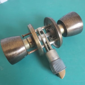 May include: An antique brass door knob assembly. It features two cone-shaped knobs, a central mechanism, and a latch. The metal components have a weathered appearance. The assembly is set against a teal background.