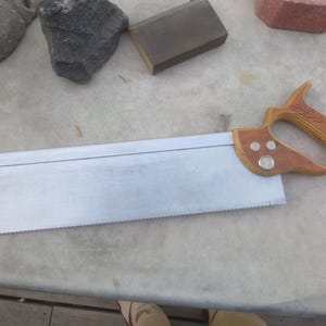 Peut inclure: Une scie à main avec une poignée en bois et une lame argentée. La poignée est marron avec un motif de grain de bois plus clair et trois rivets argentés. La lame a une série de dents le long du bord inférieur.