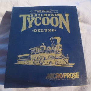 May include: A blue box with gold lettering for the game Sid Meier's Railroad Tycoon Deluxe. The box features a gold illustration of a steam locomotive.