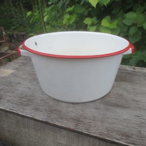 May include: A white enamelware basin with a red rim and handles. The basin is sitting on a weathered wooden surface, with greenery in the background. This vintage-style basin is suitable for various household uses.