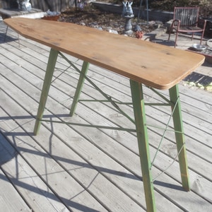 May include: A vintage ironing board featuring a long, tapered wooden top with a natural light brown finish. The base consists of four green-painted wooden legs connected by a series of thin, crisscrossing metal support wires for stability.
