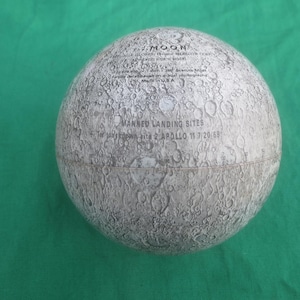 May include: A spherical model of the moon, featuring a detailed surface with craters and text. The text includes "MOON" and "MANNED LANDING SITES." The model is gray and white, resting on a green surface.