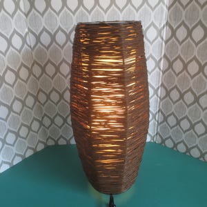 May include: A brown wicker lampshade with a light bulb inside. The lampshade is cylindrical and has a woven pattern.