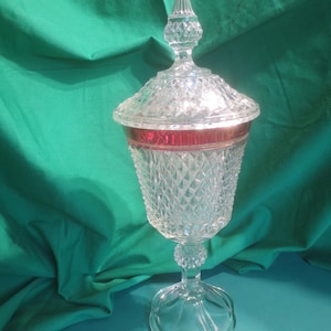 May include: A clear, lidded glass candy dish with a diamond-cut pattern. The rim features a red band. The dish sits on a pedestal base. The lid is topped with a decorative finial. The background is a solid green.