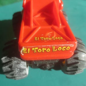 May include: A red toy monster truck with black tires. The text "El Toro Loco" is printed in yellow on the back of the truck. The truck is on a teal surface.