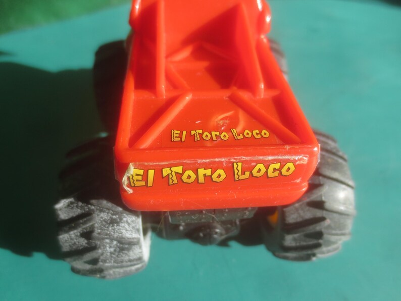 May include: A red toy monster truck with black tires. The text "El Toro Loco" is printed in yellow on the back of the truck. The truck is on a teal surface.