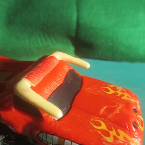 May include: A red toy monster truck with yellow flames painted on the hood and a yellow roll bar. The truck has a black windshield and a smiling mouth design. The toy is on a teal surface with a green background.