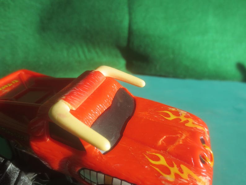 May include: A red toy monster truck with yellow flames painted on the hood and a yellow roll bar. The truck has a black windshield and a smiling mouth design. The toy is on a teal surface with a green background.