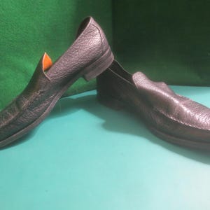 May include: A pair of black leather slip-on shoes. The shoes have a textured surface and a classic design. The soles are dark, and the interior is a light orange color. The shoes are displayed on a green and blue surface.