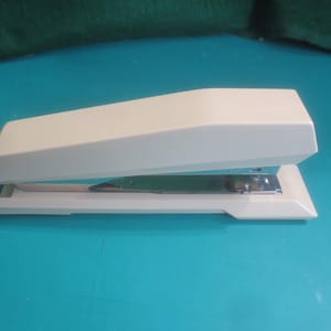 May include: A cream-colored stapler with a rectangular base and a curved top. The stapler is open, revealing the metal stapling mechanism. The background is a solid teal color.