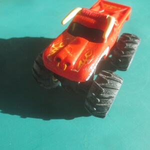 May include: A red toy monster truck with black tires and a yellow flame design on the hood. The truck has white horns and a black windshield. The toy is on a teal surface.