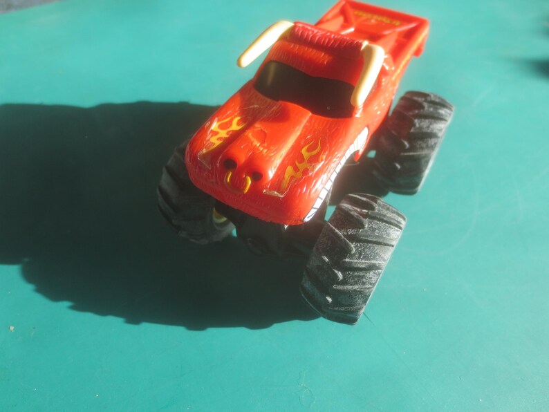 May include: A red toy monster truck with black tires and a yellow flame design on the hood. The truck has white horns and a black windshield. The toy is on a teal surface.