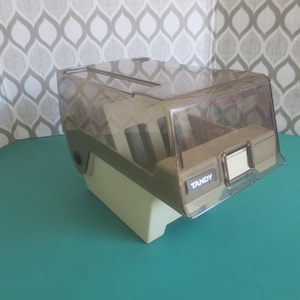 May include: A clear plastic storage box for floppy disks with a beige base and a TANDY label. The box has a hinged lid and slots for holding multiple floppy disks.