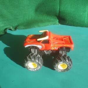 May include: A red monster truck toy with large black tires and yellow rims. The truck has a menacing grin on the front and the text "El Toro Loco" on the side. The truck has a flame design on the side.
