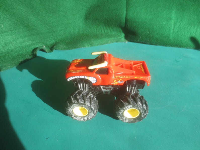 May include: A red monster truck toy with large black tires and yellow rims. The truck has a menacing grin on the front and the text "El Toro Loco" on the side. The truck has a flame design on the side.