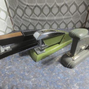Puede incluir: Se muestran tres grapadoras antiguas. Una es negra con una parte superior de madera, otra es verde oliva con detalles cromados y la tercera es de diseño gris oscuro y redondeado. Es probable que estos artículos de oficina sean de mediados del siglo XX.