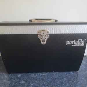 May include: Black portafile carrying case with a silver latch and a silver trim around the top. The case has the text "portafile™ by ANTHIS" printed on the front.