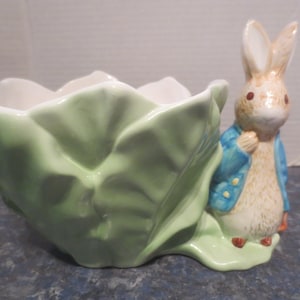 May include: A ceramic planter shaped like a cabbage with a Peter Rabbit figurine peeking out from the side. The planter is green and white with a brown rabbit figurine wearing a blue jacket.