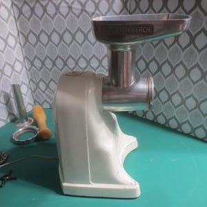 May include: White electric meat grinder with a silver metal attachment. The grinder has the words "Dayton Beach" printed on the top. The grinder is on a green surface with a wooden tamper, a metal tube, and a metal plate.