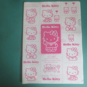 May include: A translucent plastic document folder featuring a repeating pink graphic print of Hello Kitty characters, small teddy bears, and hearts. The text Hello Kitty is printed in pink throughout the design, with a central pink rectangular panel.
