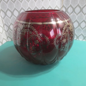 Peut inclure: Un vase rond en verre rouge rubis avec une large ouverture. Le vase présente un motif floral et de feuilles dorées autour du milieu et sur les côtés. Le vase est posé sur une surface turquoise.