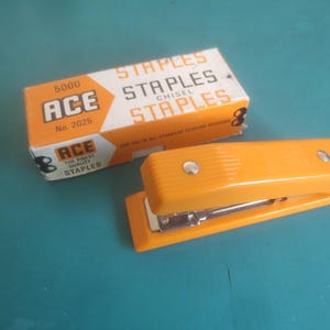 May include: An orange stapler and a box of staples. The stapler is a bright orange color with a silver metal base. The box is orange and white with the brand name "ACE" and the word "STAPLES" printed on it.