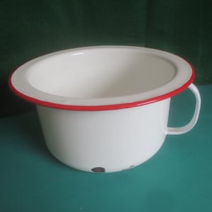 May include: White enamel chamber pot with a red rim and handle. The pot has a rounded shape and a wide opening. The handle is small and curved. The pot shows some signs of wear.