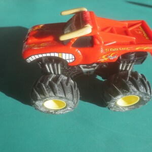 May include: A red toy monster truck with large black tires and yellow rims. The truck has a bull-like design with horns and a toothy grin. The text "El Toro Loco" is visible on the side.