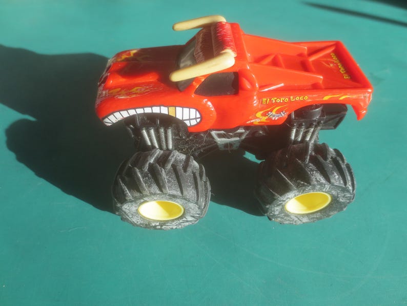 May include: A red toy monster truck with large black tires and yellow rims. The truck has a bull-like design with horns and a toothy grin. The text "El Toro Loco" is visible on the side.