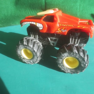 May include: A red toy monster truck with large black tires and yellow rims. The truck has a cartoonish face design with a wide grin and the text "El Toro Loco" on the side. The truck is designed for play.