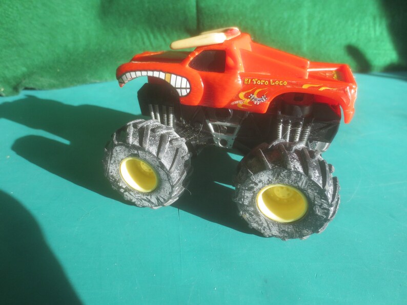 May include: A red toy monster truck with large black tires and yellow rims. The truck has a cartoonish face design with a wide grin and the text "El Toro Loco" on the side. The truck is designed for play.