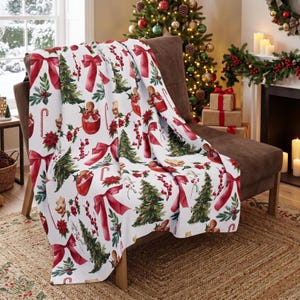 May include: A white throw blanket featuring a repeating Christmas pattern of red bows, green pine trees, gingerbread figures in mugs, candy canes, holly berries, and poinsettia flowers draped over a brown chair in a festive room.